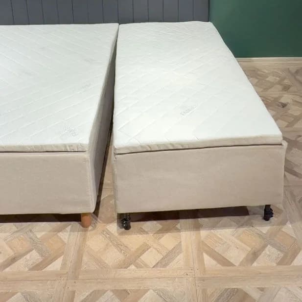 Due moduli letto completi affiancati su un pavimento in legno chiaro, separati da una stretta fessura. Ogni modulo ha il proprio rivestimento beige, il proprio materasso trapuntato e le proprie gambe.