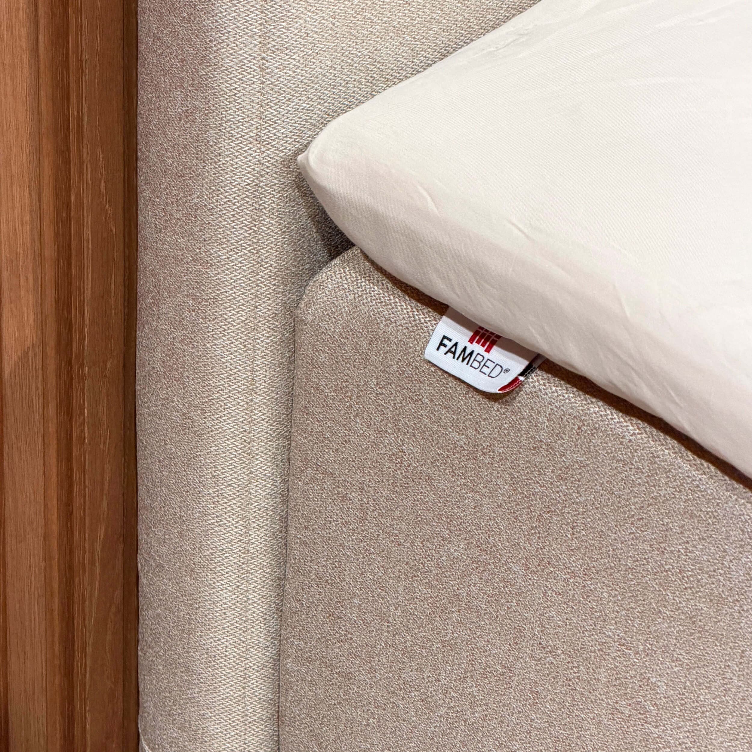 Primo piano del rivestimento beige sull'angolo del letto, con etichetta FAMBED e lenzuolo bianco sovrapposto
