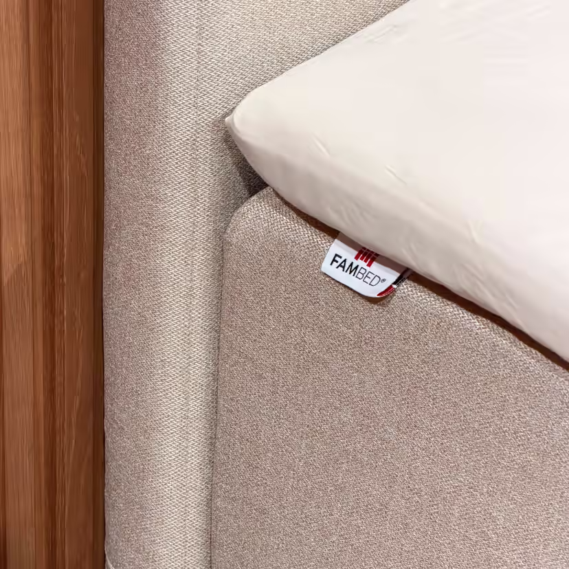Primo piano del rivestimento beige sull'angolo del letto, con etichetta FAMBED e lenzuolo bianco sovrapposto